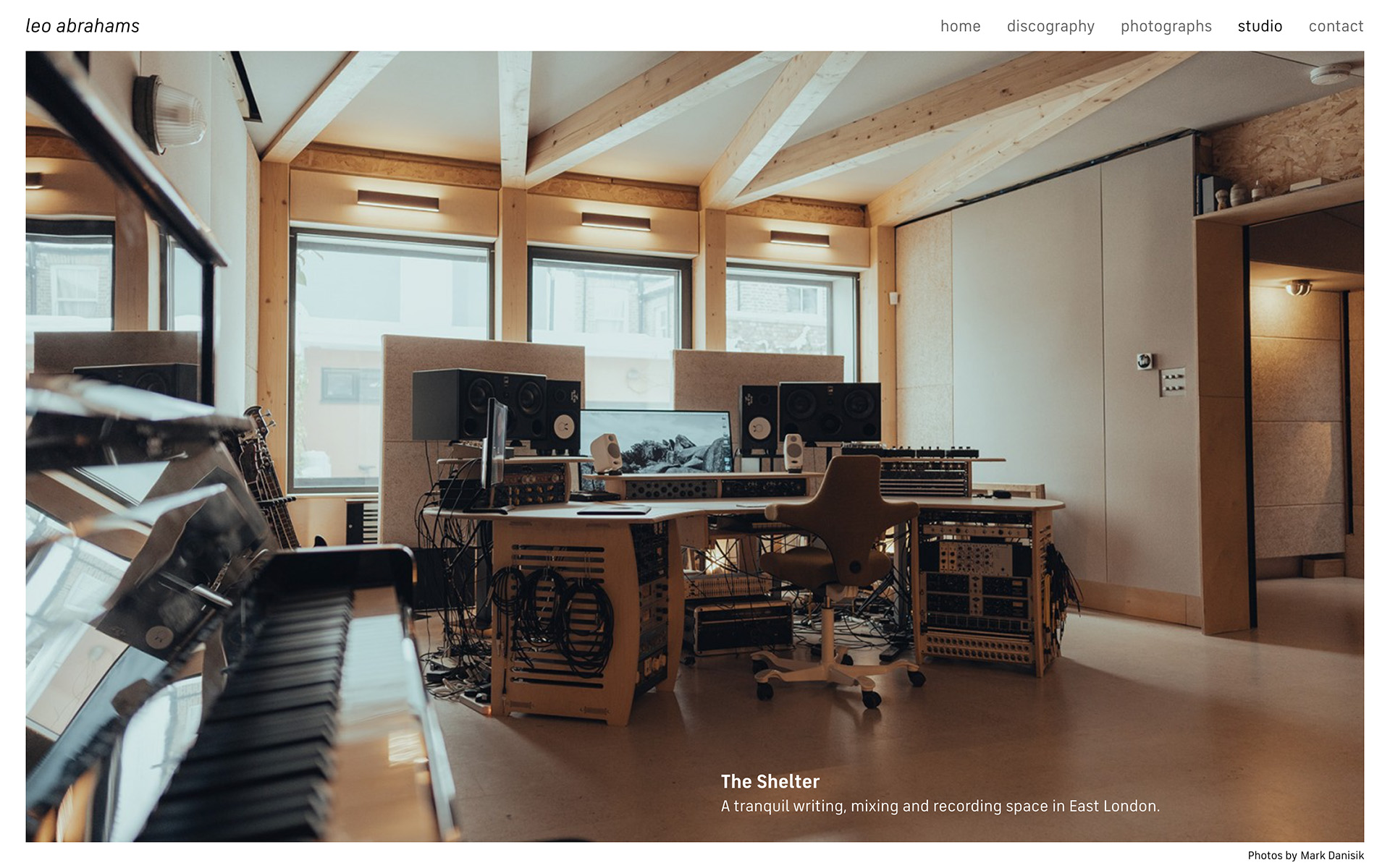 A photograph of Leo Abrahams recording studio, The Shelter. Taken from the back of the room, you can see a nice modern room with wooden beams and a minimal control centre with outboard equipment shared across two racks.